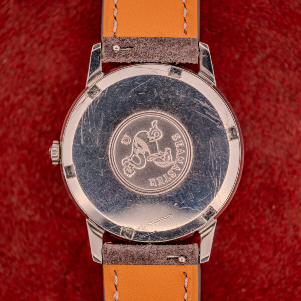 Back of a scratched Omega Seamaster Automatic Ref. 165.002 with Cal. 552 movement, shown with a tan leather strap on a red textured surface.