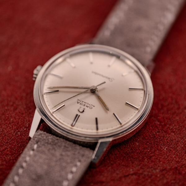 An Omega Seamaster Automatic Ref. 165.002 Cal. 552 with a grey suede strap is displayed on a red textured surface.