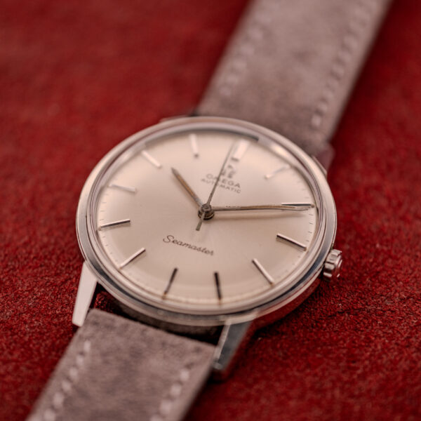 An Omega Seamaster Automatic Ref. 165.002 with Cal. 552, featuring a beige dial and grey suede strap, displayed on a red textured surface.