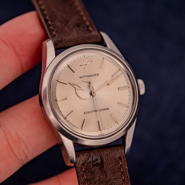 A hand holds a Wittnauer Electro-Chron "Lightning Bolt" Ref. 6150/2 Cal. 11EW c.1962, featuring a brown leather strap, silver dial, and its signature lightning bolt seconds hand.