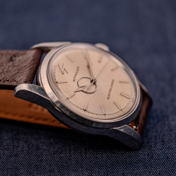 A close-up of a Wittnauer Electro-Chron "Lightning Bolt" Ref. 6150/2 Cal. 11EW c.1962 wristwatch with a brown leather strap is shown resting on a dark blue fabric background.
