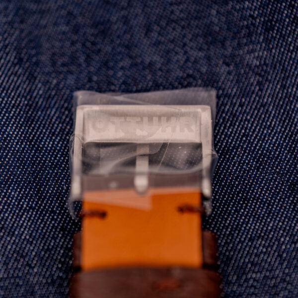 A close-up of a belt buckle covered in plastic wrap, set against denim fabric—evoking the gleaming precision of a Wittnauer Electro-Chron "Lightning Bolt" Ref. 6150/2 Cal. 11EW c.1962 timepiece.