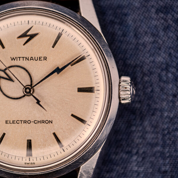 Close-up of a Wittnauer Electro-Chron "Lightning Bolt" Ref. 6150/2 Cal. 11EW c.1962 wristwatch with a silver dial, black hour markers, and distinctive lightning bolt seconds hand, displayed on blue fabric.