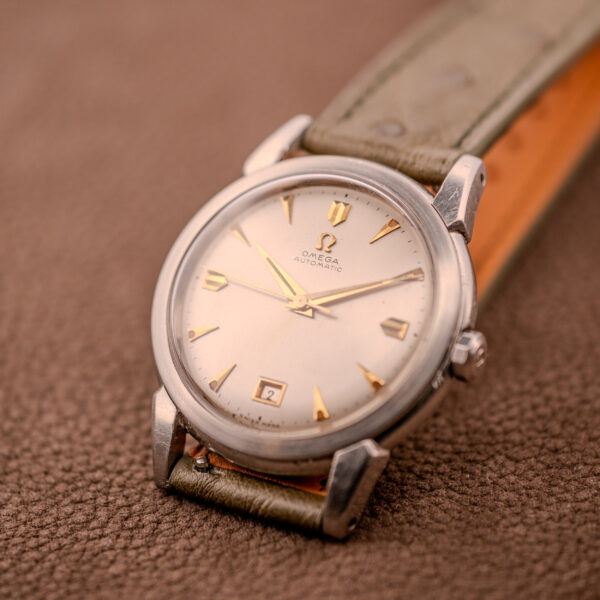 An Omega Transitional Calendar Bumper Automatic Ref. 2438-2 Cal. 353 c.1951 wristwatch with a round dial and leather strap is displayed on a brown textured surface.
