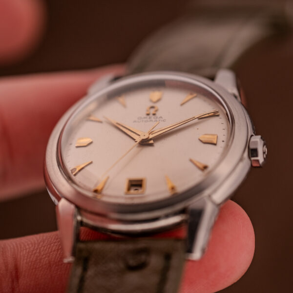 A close-up of a hand holding an Omega Transitional Calendar Bumper Automatic Ref. 2438-2 Cal. 353 c.1951 wristwatch with a leather strap and gold hour markers.