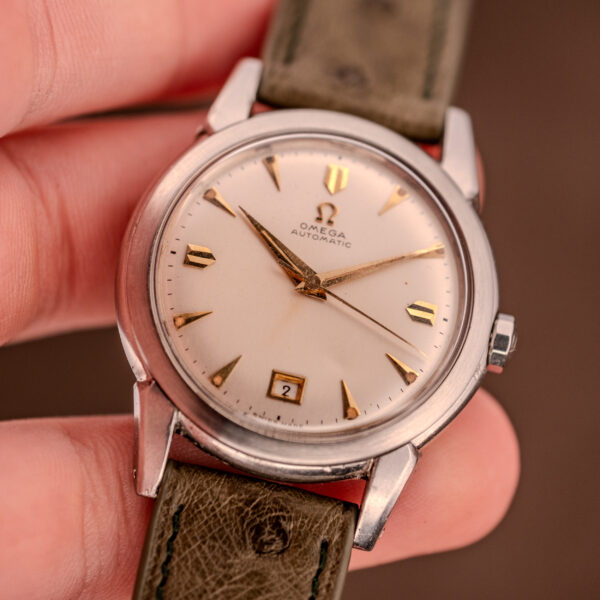 A hand holds an Omega Transitional Calendar Bumper Automatic Ref. 2438-2 Cal. 353 c.1951 wristwatch with a cream dial, gold hour markers, and a green leather strap.