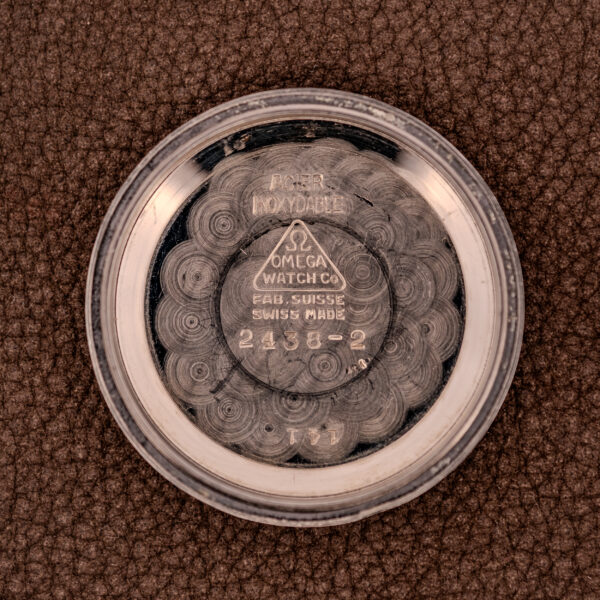 Round metal case back with Omega Watch Co. markings and numbers, featuring the Omega Transitional Calendar Bumper Automatic Ref. 2438-2 Cal. 353 c.1951, on a textured brown surface.