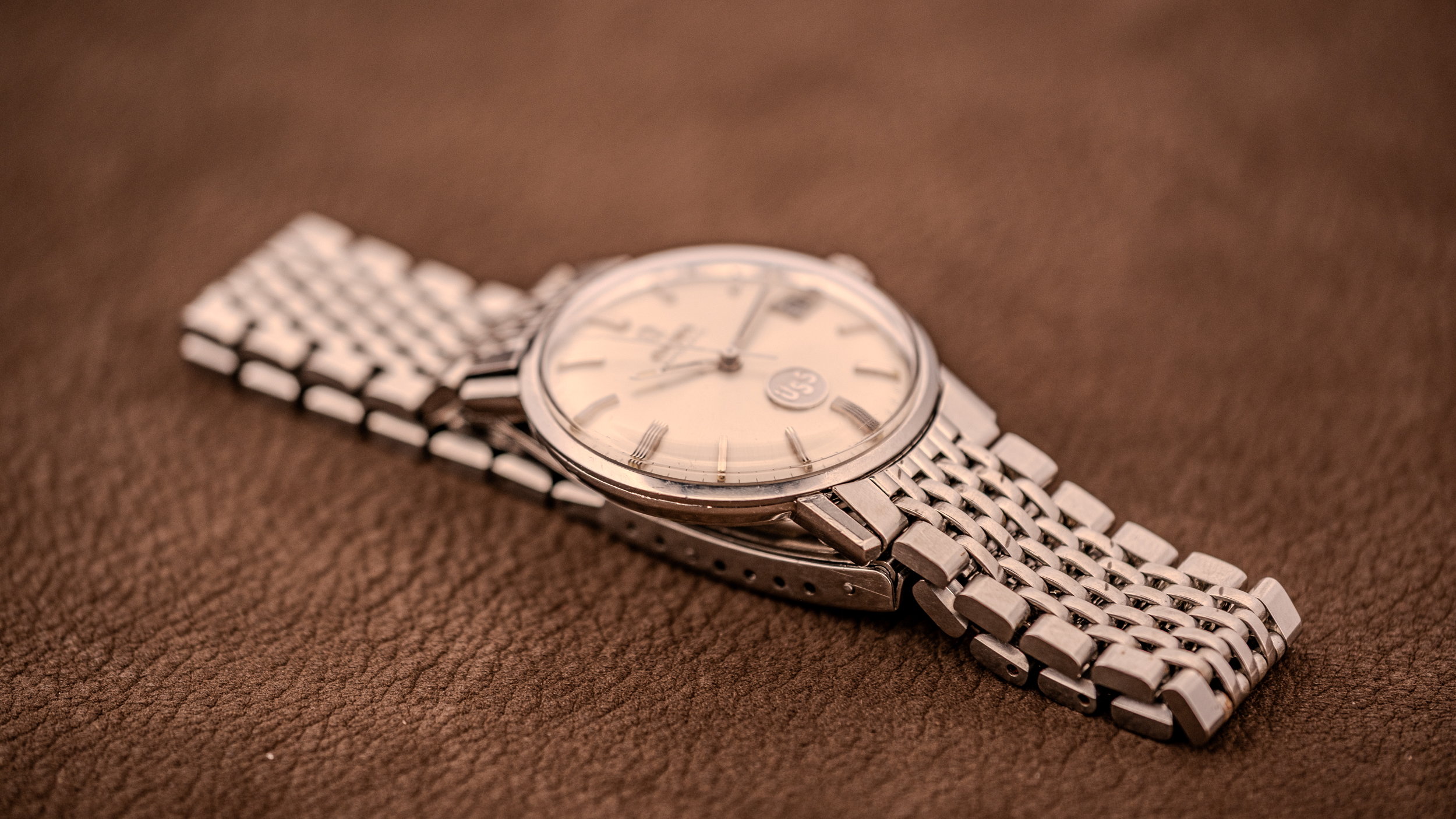 A vintage Omega Seamaster DeVille wristwatch with a silver metal band rests elegantly on a brown textured surface.