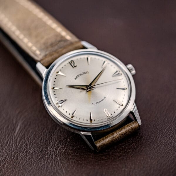The Hamilton Automatic Silver Sunburst Micro-Rotor Cal. 666 wristwatch with a tan leather strap rests elegantly on a brown surface.