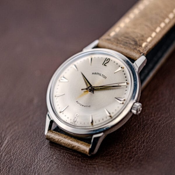 Close-up of a Hamilton Automatic Silver Sunburst Micro-Rotor Cal. 666 wristwatch with a brown leather strap and silver sunburst dial on a brown surface.