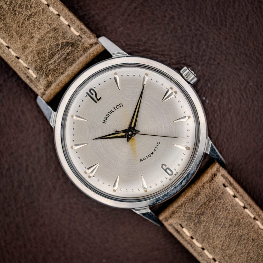 A Hamilton Automatic Silver Sunburst Micro-Rotor Cal. 666 wristwatch with a silver sunburst dial and tan leather strap rests on a brown leather surface.