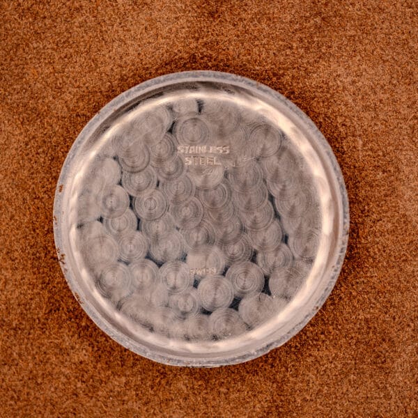 A round, scratched stainless steel Jaeger Tri-Compax Chronograph Ref. 22522 Cal. UG 285 watch back rests on a brown textured surface, viewed from above.