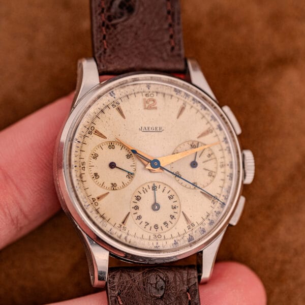 A Jaeger Tri-Compax Chronograph Ref. 22522 with stainless steel case, caliber UG 285, and brown leather strap is held between two fingers against a tan background.