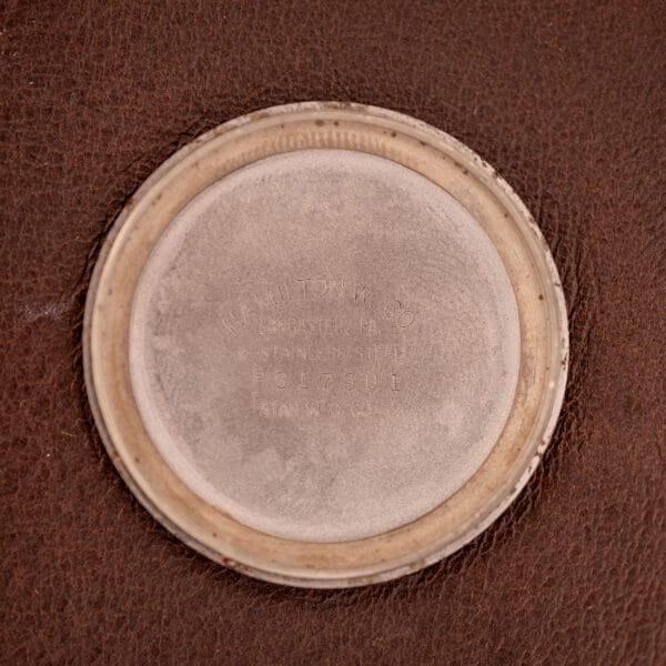 A round metal watch backplate with engraved text rests on brown leather, highlighting the refined craftsmanship of the Hamilton Automatic Silver Sunburst Micro-Rotor Cal. 666.