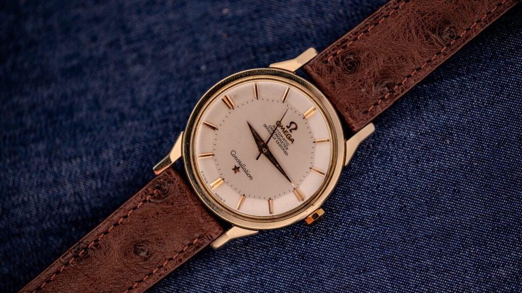 A gold Omega Constellation wristwatch with a classic pie-pan dial and a brown leather strap displayed on blue denim fabric.