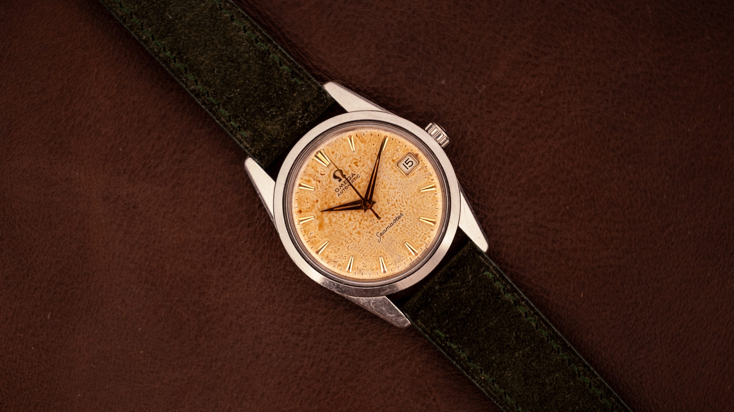 A wristwatch with a gold textured face and dark green strap on a brown leather surface, ideal for those seeking small watches for men.