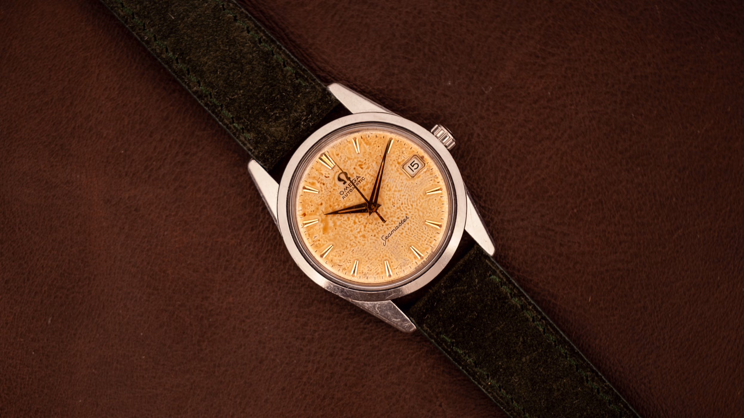 A wristwatch with a gold textured face and dark green strap on a brown leather surface, ideal for those seeking small watches for men.