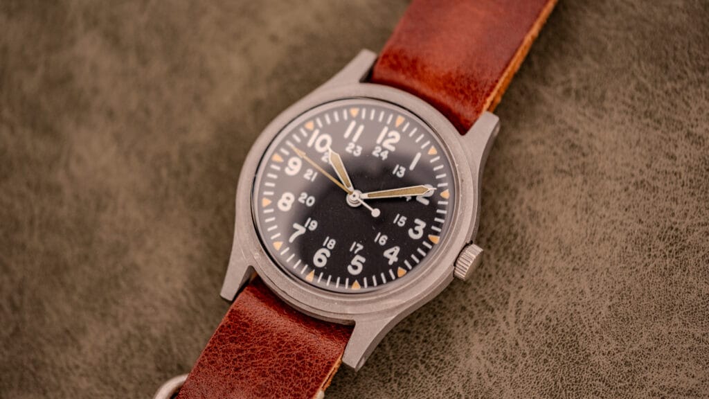A vintage field watch featuring a brown leather strap and a black dial with white numbers on a textured surface, inspired by classic GG-W-113 vintage military watches.