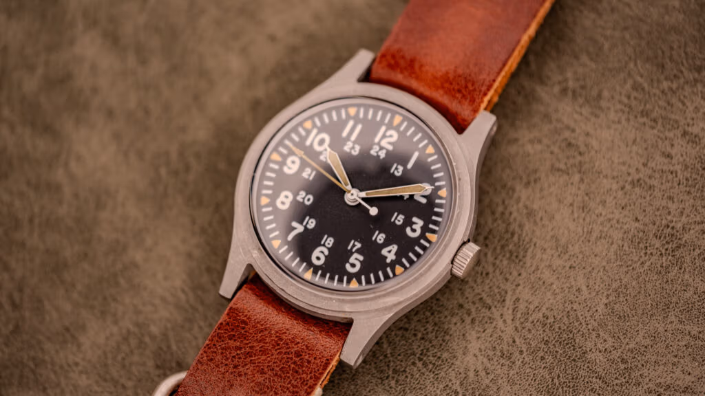A vintage field watch featuring a brown leather strap and a black dial with white numbers on a textured surface, inspired by classic GG-W-113 vintage military watches.