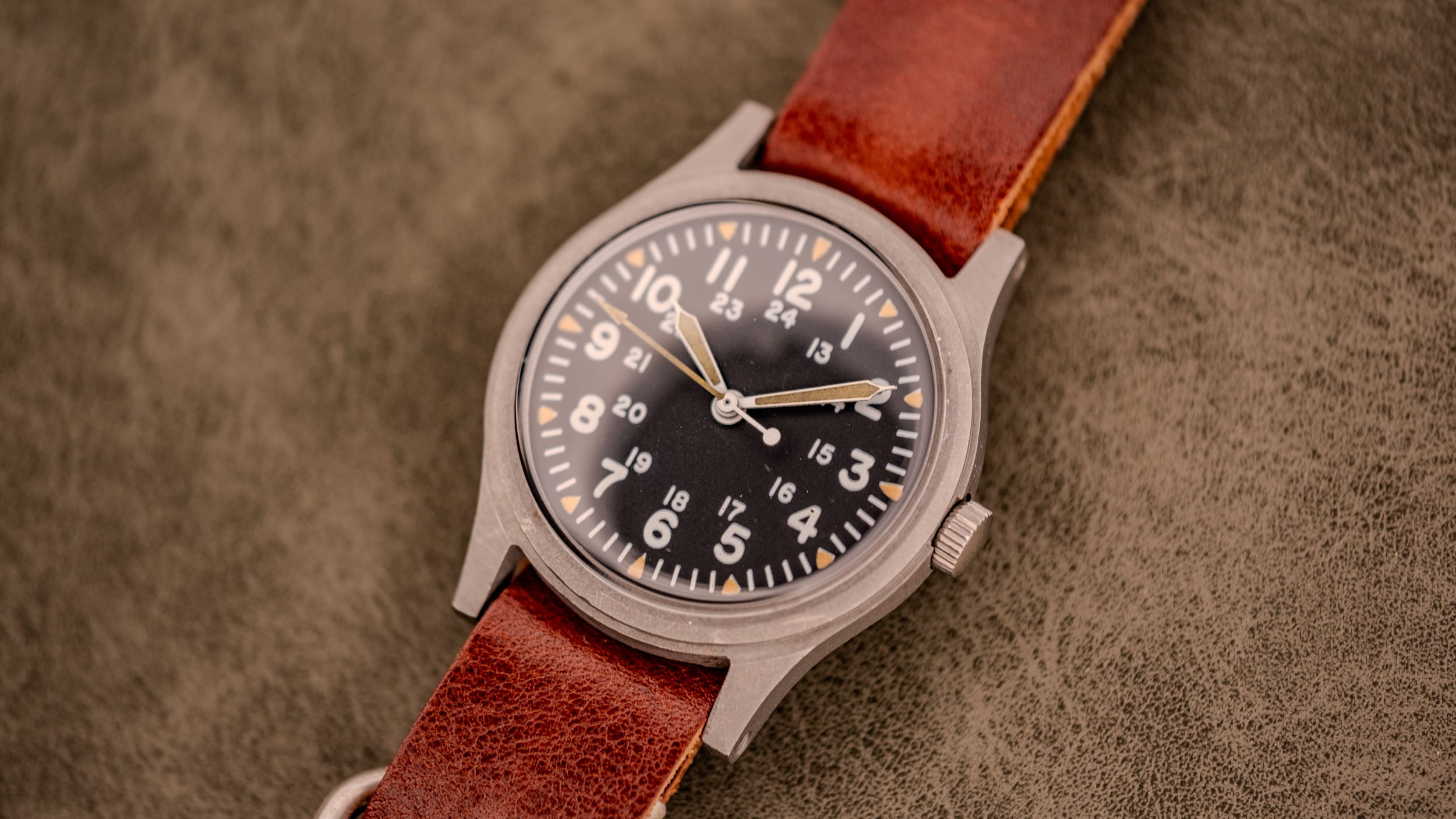 A vintage field watch featuring a brown leather strap and a black dial with white numbers on a textured surface, inspired by classic GG-W-113 vintage military watches.