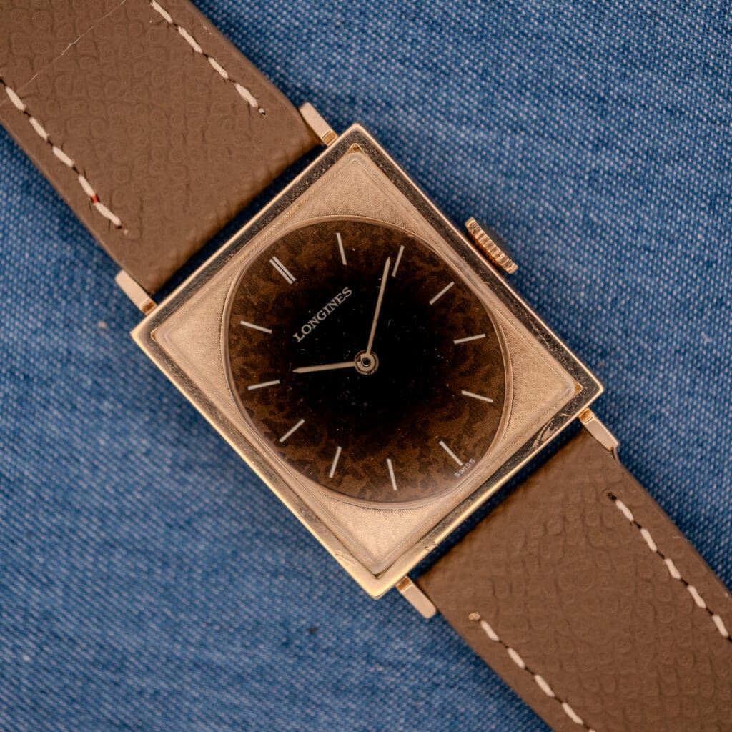 Vintage Longines Tank watch ref 3217-428 with factory Leopard dial aged into speckled tortoiseshell patina on a mud grey textured leather strap, top-down on denim
