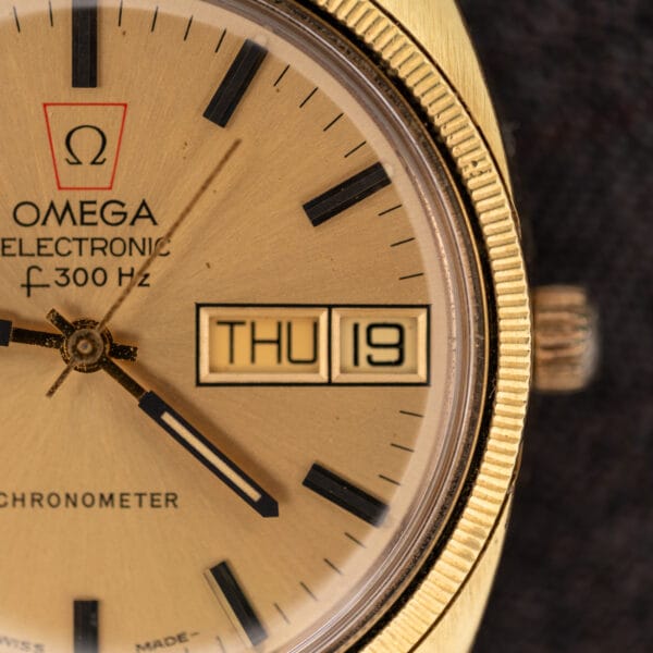 Macro of the Omega Electronic F-300 Hz champagne dial showing the red Omega cartouche, day-date window at three, and gold stick markers with black inlays