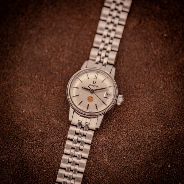 Vintage Omega Ladymatic ref. 566.002 with steel beads-of-rice bracelet laid across textured brown leather backdrop