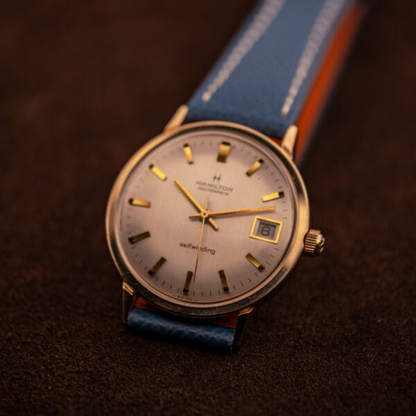 Hamilton Masterpiece automatic on blue Saffiano strap photographed top-down on tobacco suede with warm gold case