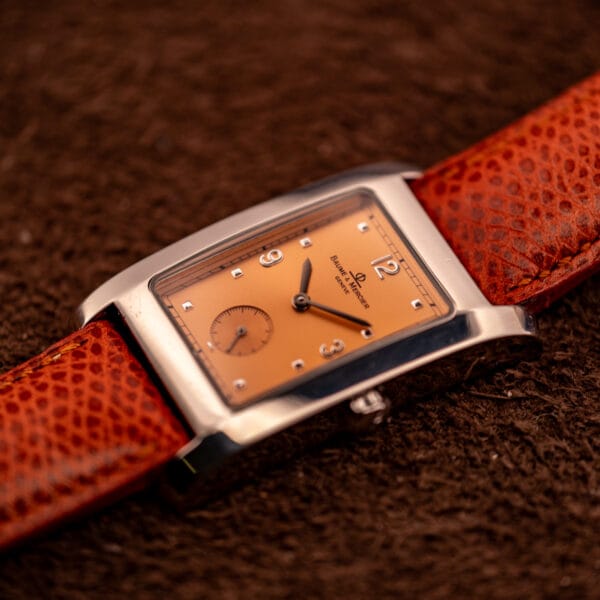 Baume & Mercier Hampton MV045063 three-quarter case profile against textured ground, showing the convex bezel and copper salmon dial