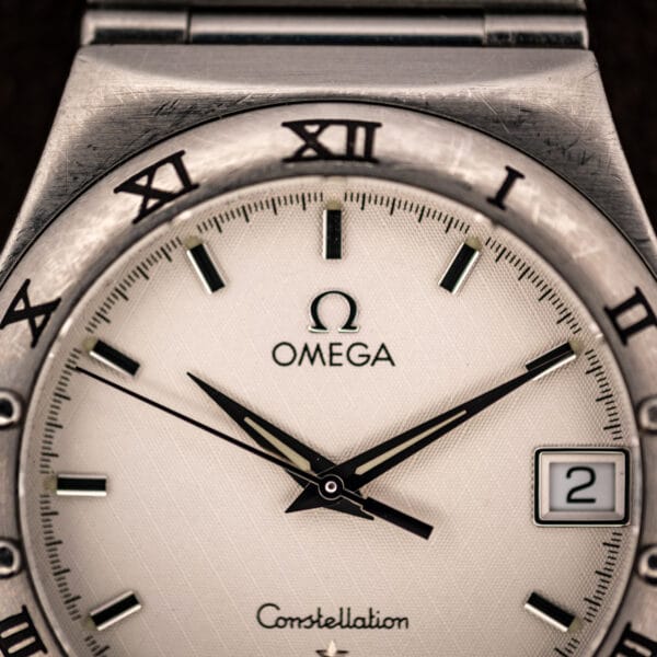 Omega Constellation 1512.30 dial close-up showing applied Omega logo, baton hour markers, and faceted dauphine hands over the woven texture