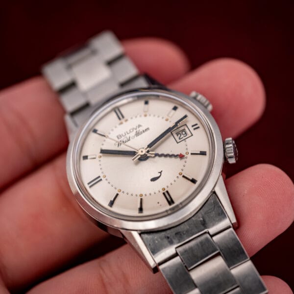 The Bulova Wrist Alarm 773 in hand under warm light, the serpentine red alarm hand crossing the dial