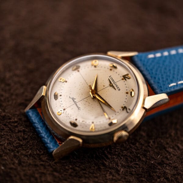 Longines crosshair dial catching low light, applied faceted markers above a blue leather strap with tan lining