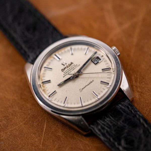 Omega Seamaster 168.022 photographed at an angle against a tan leather backdrop, date window at three and foxing visible along the upper dial.
