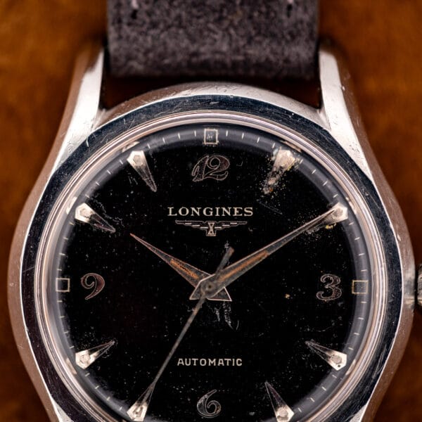 Longines 6536-1 black dial macro showing winged hourglass logo, scrolled Arabic 12 and 3, aged dauphine hands