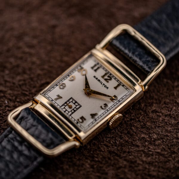 Hamilton Wilshire rectangular gold filled case at an angle, fluted crown and articulated loop lugs holding a black leather strap