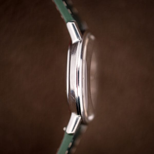Macro side view of the polished case flank, domed acrylic crystal, and green strap of the Seamaster De Ville.