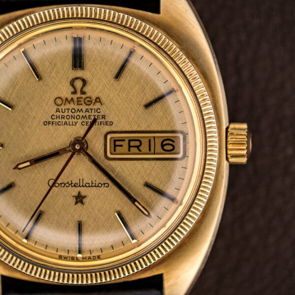 Macro of the Omega Constellation dial showing Automatic Chronometer Officially Certified text, linen texture, and the day-date aperture at three.