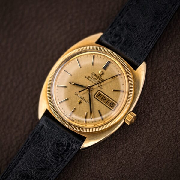 Angled view of the gold-capped Omega Constellation cushion case, fluted coin-edge bezel, and Omega-signed crown catching the light.