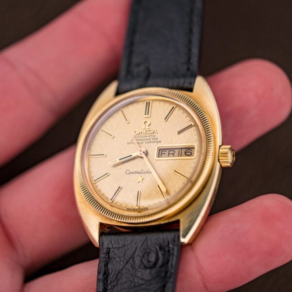 Vintage Omega day-date resting between fingers, showing the gold case proportions, fluted bezel, and black ostrich strap.