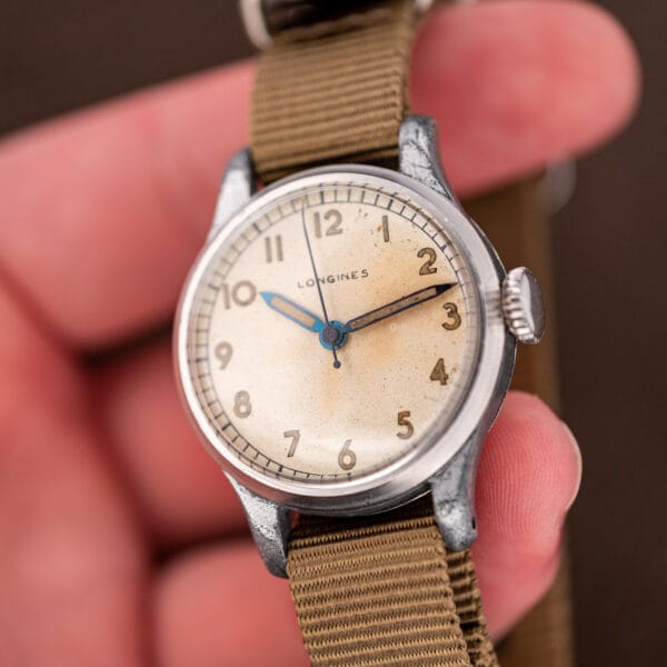Longines military-style field watch in hand, blued-steel sweep hand crossing an aged cream dial