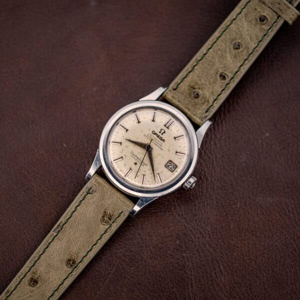 Vintage Omega Constellation 2943-2 watch laid flat on olive ostrich leather over a brown hide backdrop