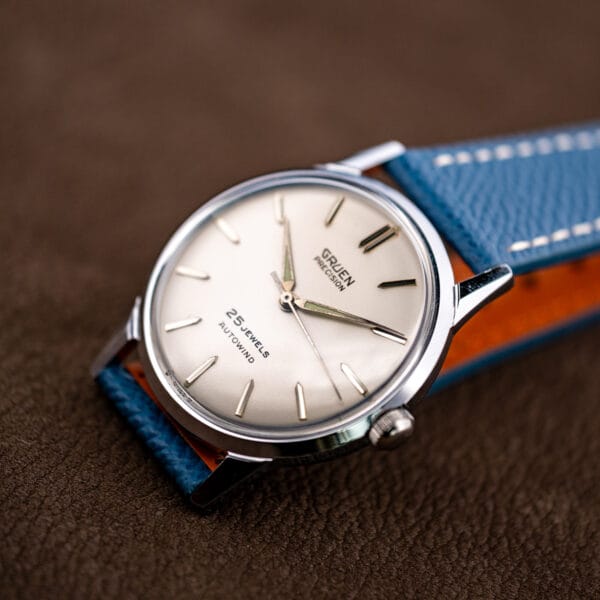 Angled beauty shot of the silvered Gruen dial, faceted baton markers and dauphine hands over blue leather.