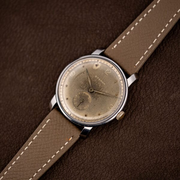 Eterna dress watch on taupe grained leather strap with contrast stitching, shot at an angle on brown leather