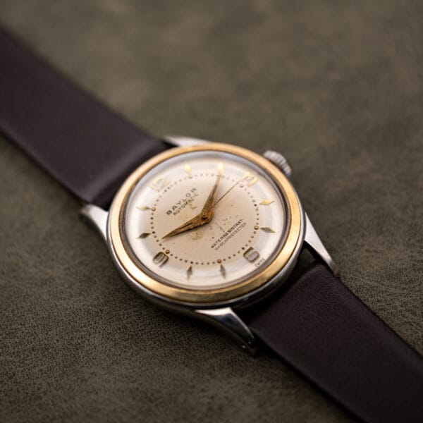 Vintage Baylor watch resting on leather, showing case profile, yellow bezel and faceted stainless steel lugs