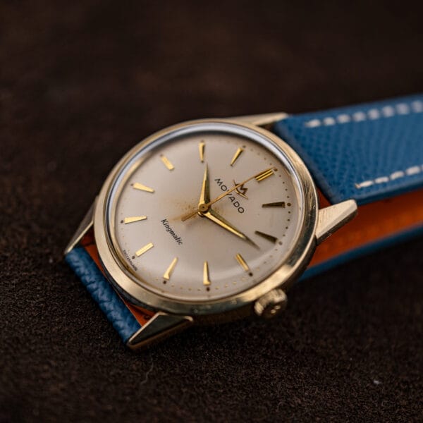 Movado Kingmatic at an angle with the blue strap curled to show its orange leather lining