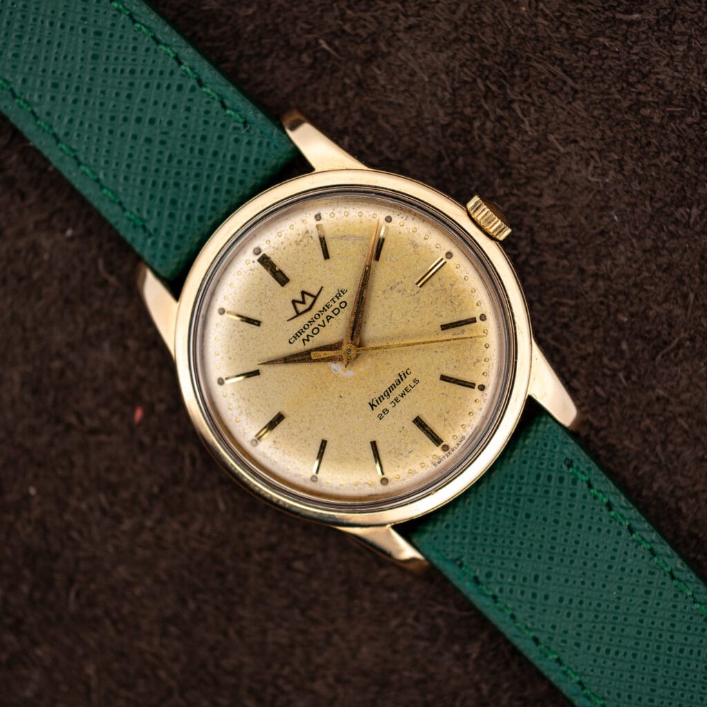 Movado Chronometre Kingmatic on green saffiano strap showing champagne dial, gold capped case and faceted dauphine hands