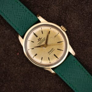 Movado Chronometre Kingmatic on green saffiano strap showing champagne dial, gold capped case and faceted dauphine hands