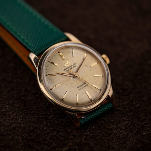 Movado Kingmatic angled on brown fabric with fluted gold crown, faceted lugs and green saffiano strap