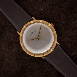Girard-Perregaux silver sunburst dial, two gold-toned hands, and a textured bezel with twelve polished notches, shown on suede