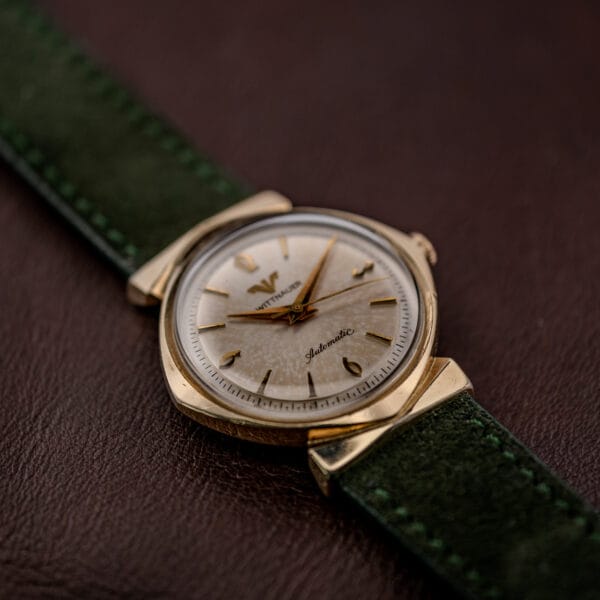 Angled view of a silver patina dial with gold tone bezel and forest green suede strap